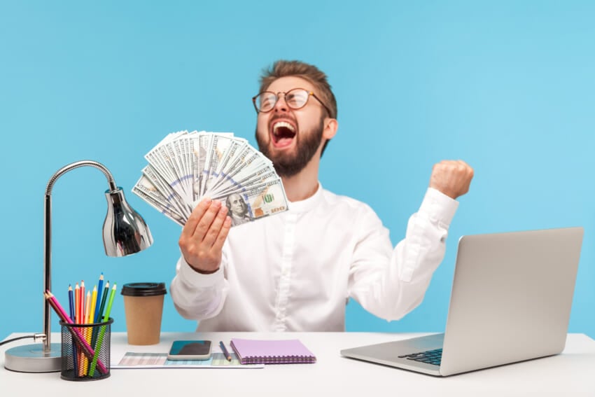 Extremely-happy-satisfied-man-office-worker-screaming-holding-in-hand-big-sum-of-dollars-winning-jackpot-at-lottery-sitting-at-workplace-with-laptop-indoor-studio-shot-isolated-on-blue-background.jpg
