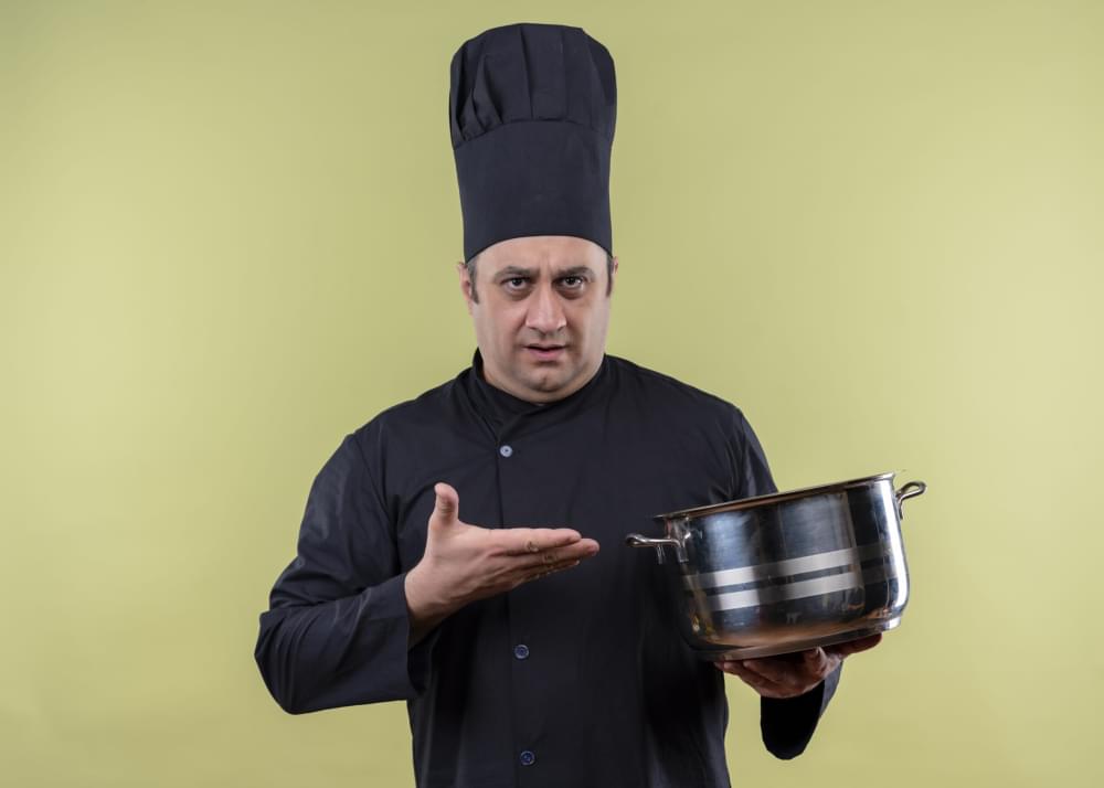 Male-chef-cook-wearing-black-uniform-and-cook-hat-demonstrating-saucepan-with-arm-looking-confused-standing-over-green-background.jpg